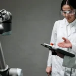 Researchers in lab coats and safety glasses engaging with a robotic arm in a lab setting.