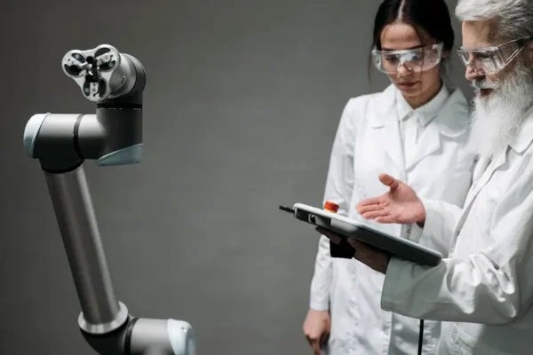 Researchers in lab coats and safety glasses engaging with a robotic arm in a lab setting.