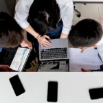 Three colleagues working together on laptops at an office desk with documents.