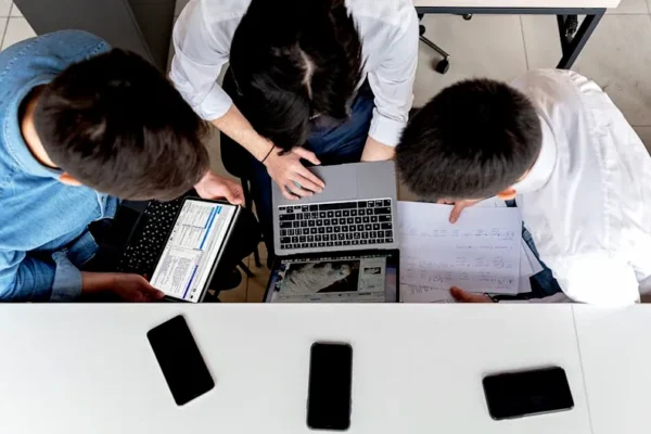 Three colleagues working together on laptops at an office desk with documents.