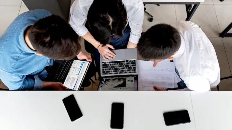 Three colleagues working together on laptops at an office desk with documents.